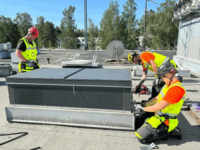 Kolme rakennusalan ammattilaista asentamassa kerrostalon katolla kirkkaassa säässä.